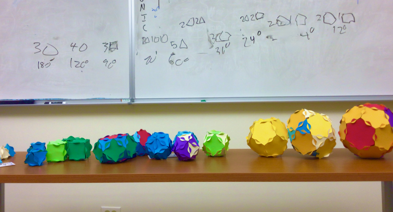 many paper polyhedra, all in line. They are sorted by angle defect, which happens to correspond to sorting by size